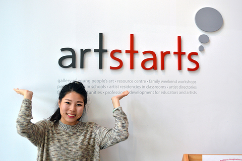 An ArtStarts volunteer stands below the ArtStarts logo mounted to the wall. The person's hands are raised up to logo
