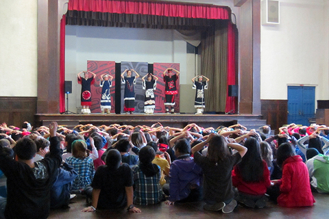 Artists on stage in a school auditorium as young learners sit on the floor with their hands on their heads mirroring the perform